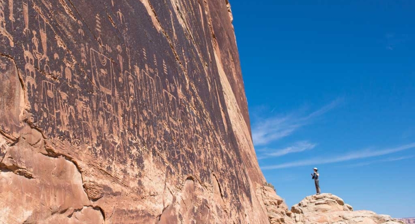 A person stands beside a tall canyon wall, looking up at ancient markings. 